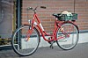 Red bicycle with a rear basket parked beside a brick wall on a sidewalk.
