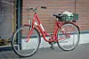 Red bicycle with a rear basket parked beside a brick wall on a sidewalk.