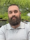 A man with a beard wearing a light gray collared shirt stands outdoors.