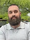 A man with a beard wearing a light gray collared shirt stands outdoors.