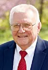 Close-up portrait of an older man with white hair, glasses, and a red tie.