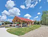 Wooden pavilion with red roof in a park next to a paved path under a blue sky.