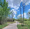 A curved paved path runs through a park with trees, grass, and a partly cloudy blue sky.