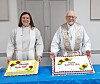 Two clergy members stand behind two decorated cakes on a blue table indoors.