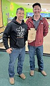 Two men posing in a bowling alley, one holding a wooden plaque-shaped award.