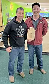 Two men posing in a bowling alley, one holding a wooden plaque-shaped award.