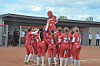 A group of players in red uniforms huddle on a dirt softball field near a dugout.