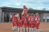 A group of players in red uniforms huddle on a dirt softball field near a dugout.