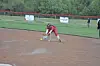 Softball player in red uniform crouches to field a yellow ball on a dirt infield.