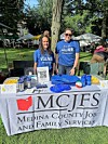 Two people behind an information table with MCJFS signage, flyers, and promotional items outdoors.