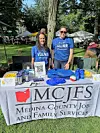 Two people behind an information table with MCJFS signage, flyers, and promotional items outdoors.