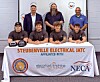 Group posing behind a Steubenville Electrical IATC banner with caps and hard hats at an indoor event.