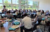 People seated at tables in a bright room with large windows, eating and talking.