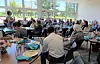 People seated at tables in a bright room with large windows, eating and talking.
