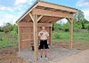 Person standing inside a small wooden shelter with a pitched roof on a gravel base.