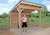 Person standing inside a small wooden shelter with a pitched roof on a gravel base.