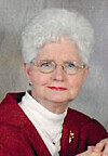 Portrait of an older woman with white hair, glasses, and a red jacket against a neutral background.