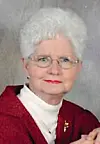Portrait of an older woman with white hair, glasses, and a red jacket against a neutral background.