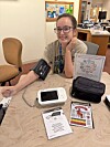 Woman seated at a table with blood pressure monitor, pamphlets, and a black bag in a clinic-like room.