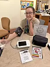 Woman seated at a table with blood pressure monitor, pamphlets, and a black bag in a clinic-like room.
