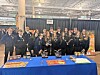A group stands behind blue-covered tables displaying brochures and packaged items at an indoor indoor event.