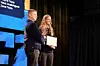 Two people on stage during an award presentation, one holding a framed plaque near a large screen.