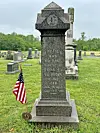 Weathered gravestone with an American flag in a cemetery with several other headstones.