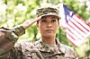 A soldier in uniform salutes outdoors with a blurred American flag behind.