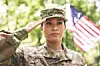 A soldier in uniform salutes outdoors with a blurred American flag behind.