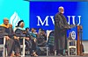 Speaker at podium addresses seated faculty on a graduation stage with a blue backdrop.