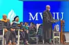 Speaker at podium addresses seated faculty on a graduation stage with a blue backdrop.