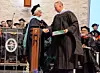 Two people in academic robes shake hands during a graduation or academic ceremony on a stage.