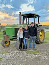 Three people standing next to a green John Deere tractor on gravel at sunset.