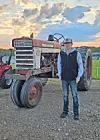 Man standing next to an old tractor on gravel at sunset.