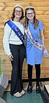 Two young women wearing tiaras and sashes stand together indoors in front of a wooden wall.