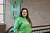 Woman in a green shirt standing near a large green H on a brick wall.