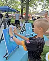 A child paints a blue canvas at an outdoor art event with other easels and tents nearby.