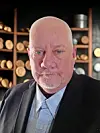 Bald man in a dark jacket standing indoors in front of shelves with dishes and containers.