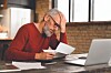 Older man in a red sweater reading papers beside an open laptop at a desk.