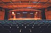 Empty theater auditorium with rows of seats facing a wooden stage under orange lighting.