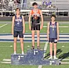 Three medal-winning athletes stand on a podium at a track stadium, with first, second and third place marked.