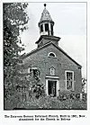 Black-and-white photo of a brick church with a steeple and overgrown plants in front.