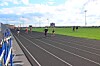 Runners on a track beside a grassy field and stadium seating.