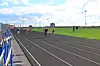 Runners on a track beside a grassy field and stadium seating.
