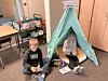 Two children sit inside a small green teepee tent in a room with tables, cabinets, and toys.