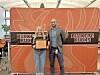 Two people stand before a Buckeye Bucks backdrop, holding a framed plaque at an indoor event.
