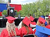 Graduates in red caps and gowns sit facing an outdoor stage with a speaker at a podium.