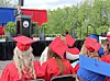 Graduates in red caps and gowns sit facing an outdoor stage with a speaker at a podium.