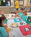 Children in a classroom work on colorful art projects at a table with craft supplies.
