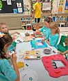 Children in a classroom work on colorful art projects at a table with craft supplies.